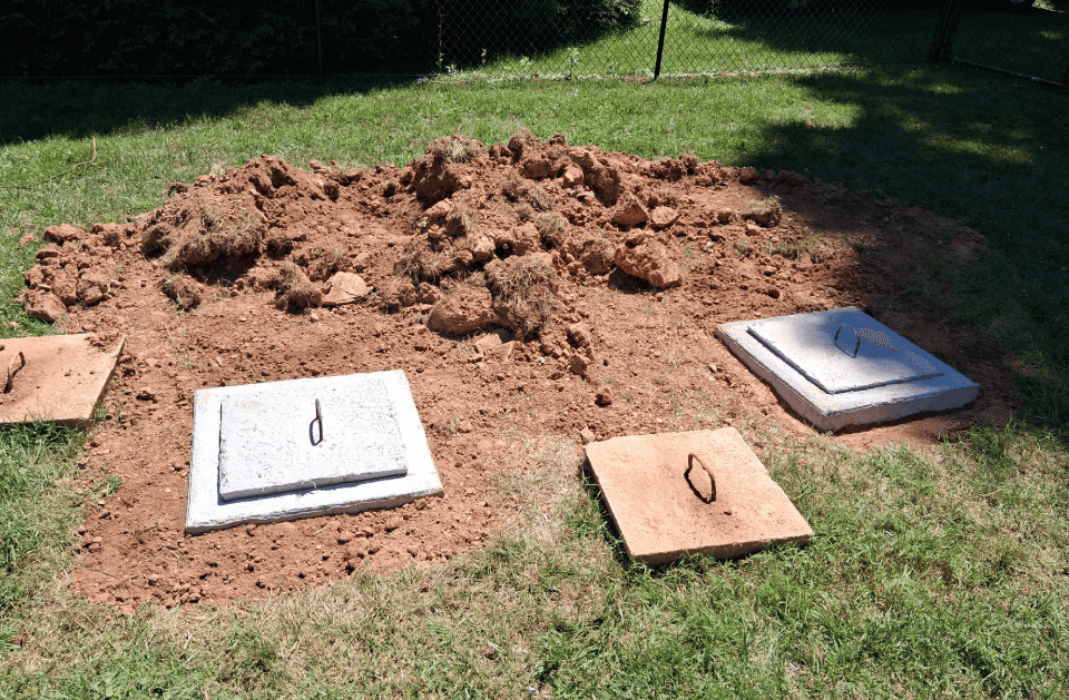 Concrete septic tank lids exposed at grade in a backyard after excavation, soil neatly piled to the side